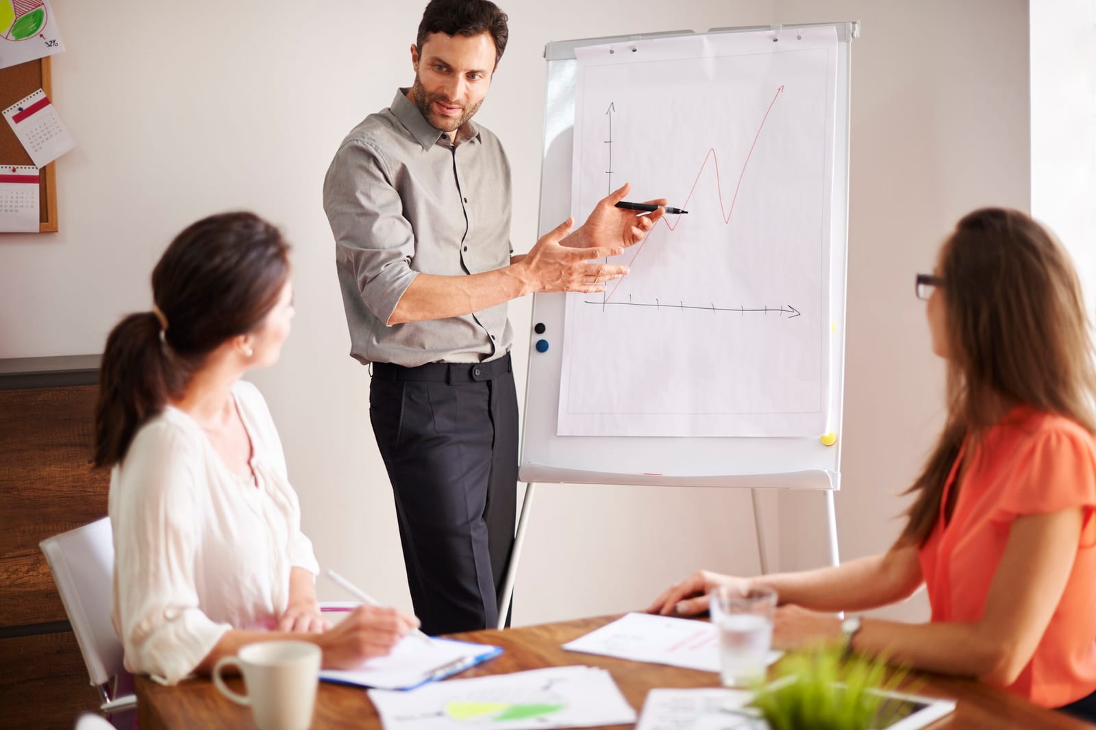 Instrutor explica um gráfico em flipchart para duas participantes sentadas à mesa.